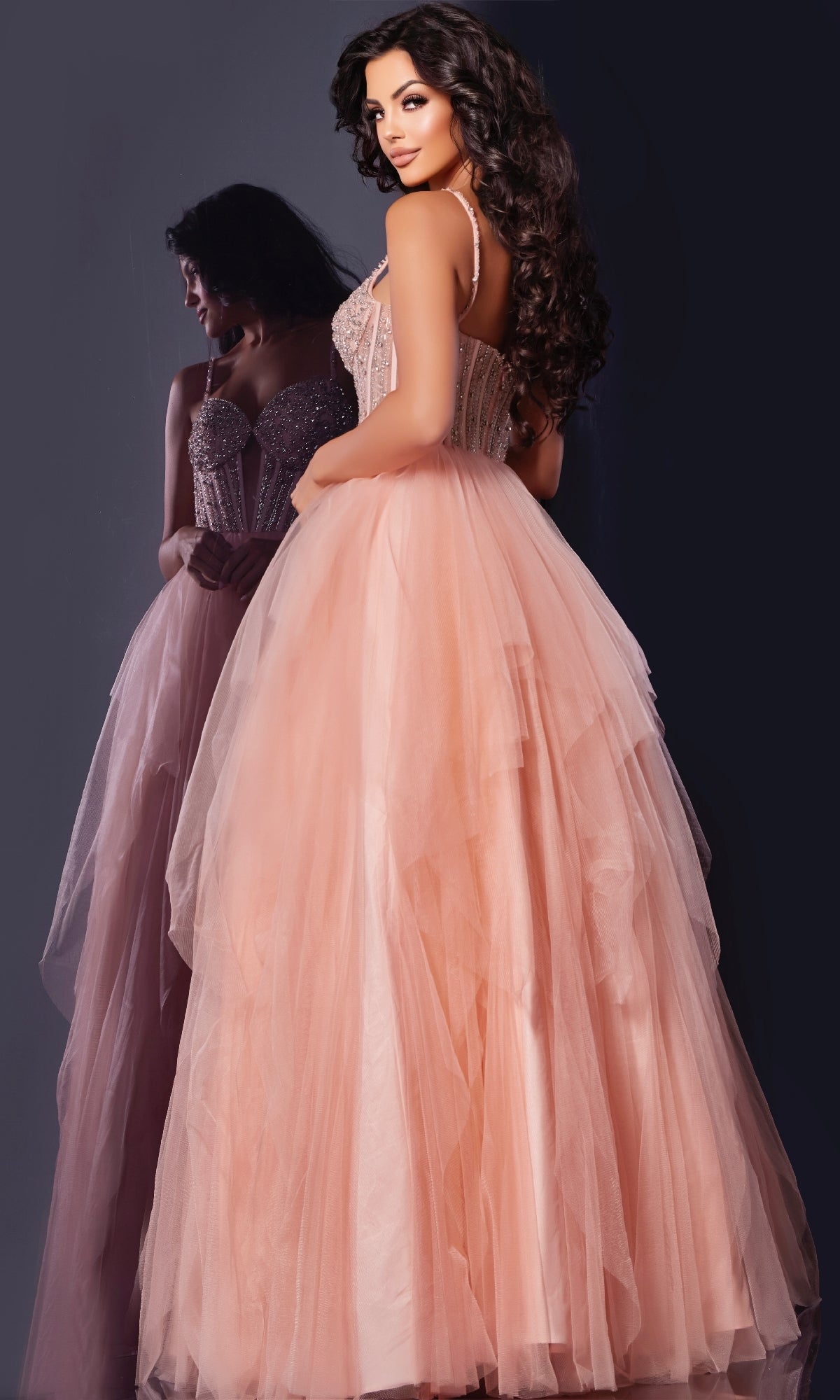 A woman models the Jovani 43254 long prom dress in blush pink, featuring an embellished corset bodice, as she stands before a mirror showcasing the gown’s beautiful back and her shimmering reflection.